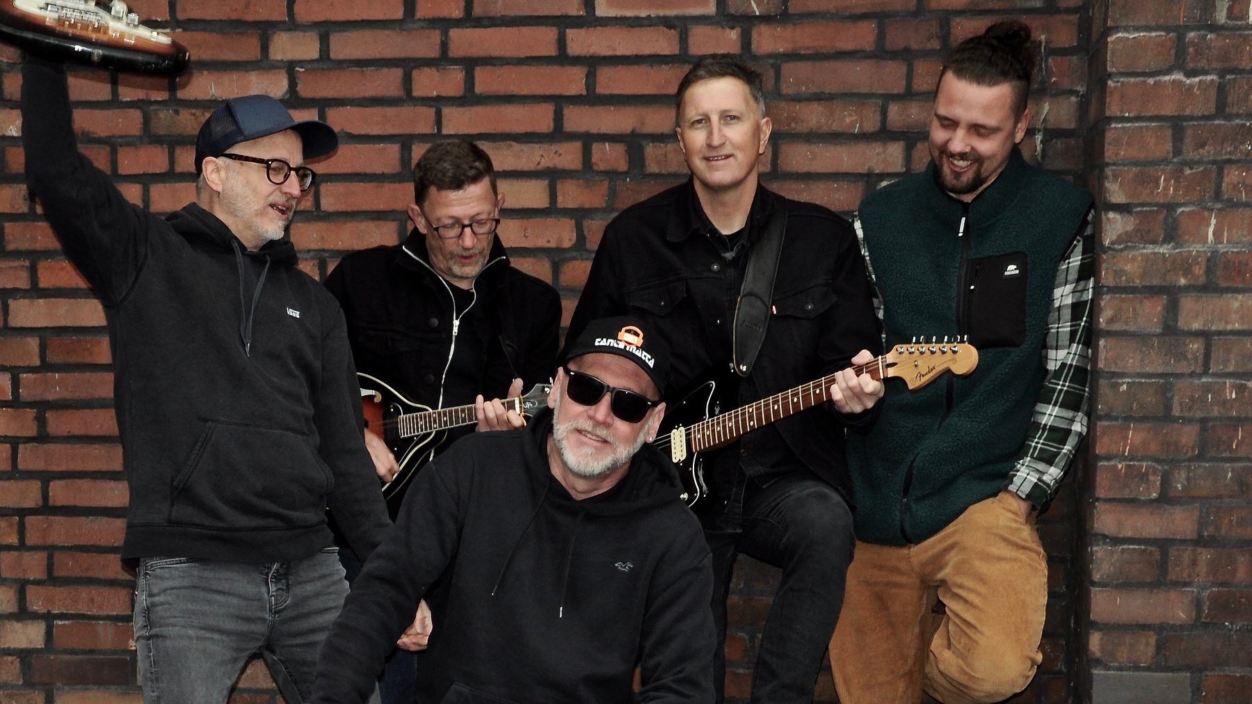 Tante Matta gut gelaunt mit Gitarren vor einer Backsteinmauer, einer reckt den Arm in die Luft